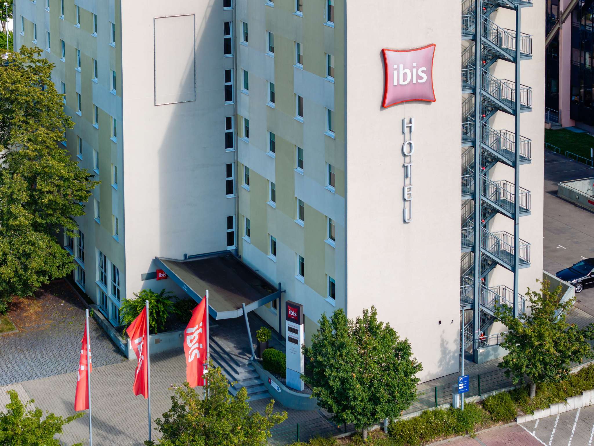 Facade of an ibis hotel with a beige/green facade, large red ibis sign, and external metal staircase on the right side.