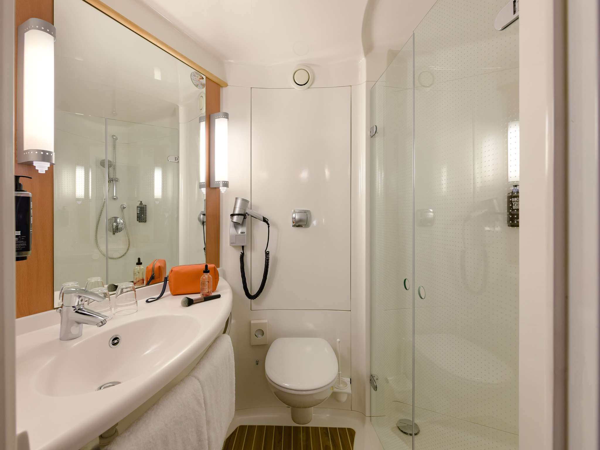 Compact bathroom with white sink, large mirror, vertical lights, toilet, and glass shower; orange bag and toiletries on the counter.