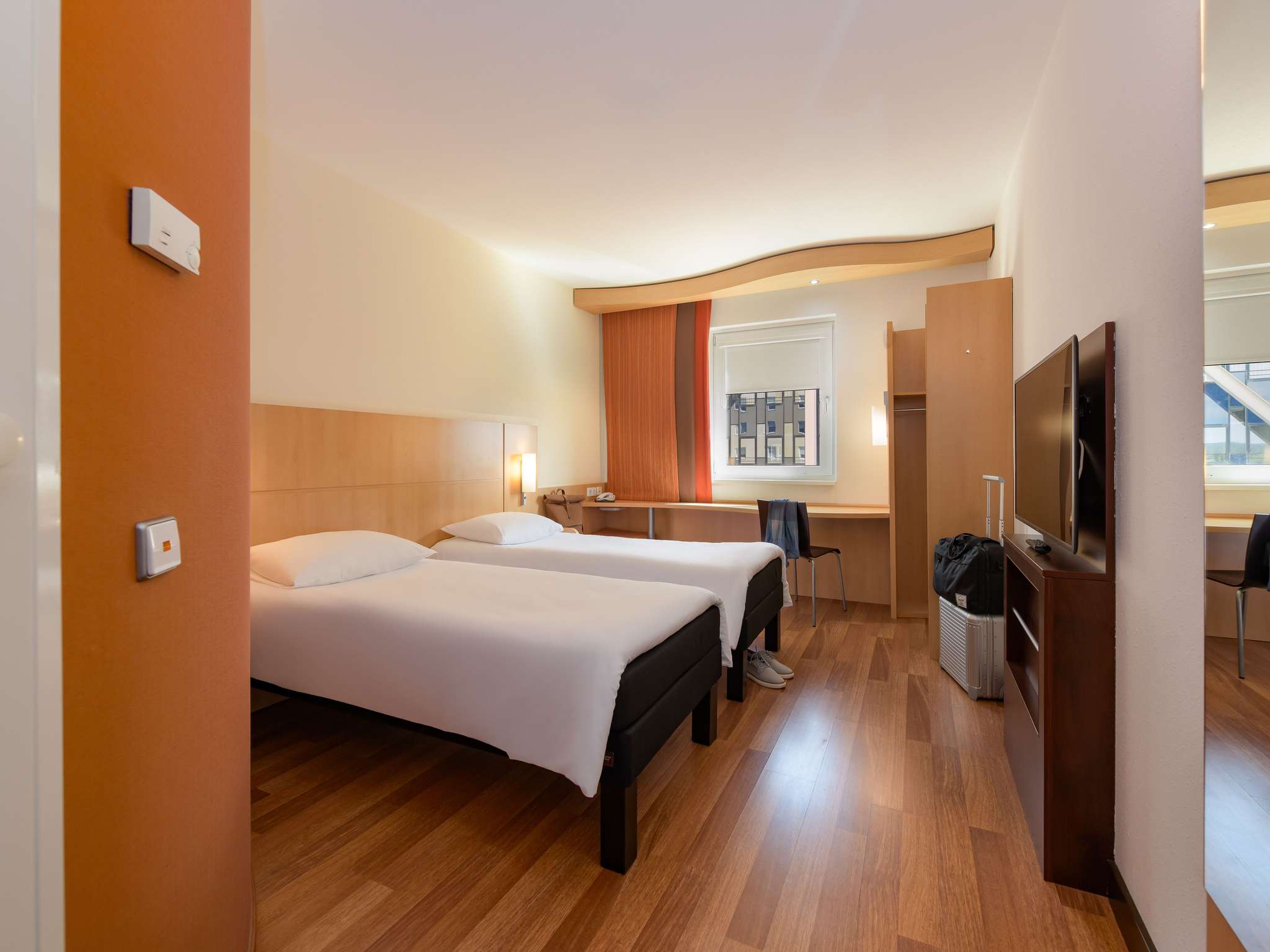 Hotel room with two single beds, wooden floor, orange accent wall, and a desk by a window with orange curtains. TV and open wardrobe visible.